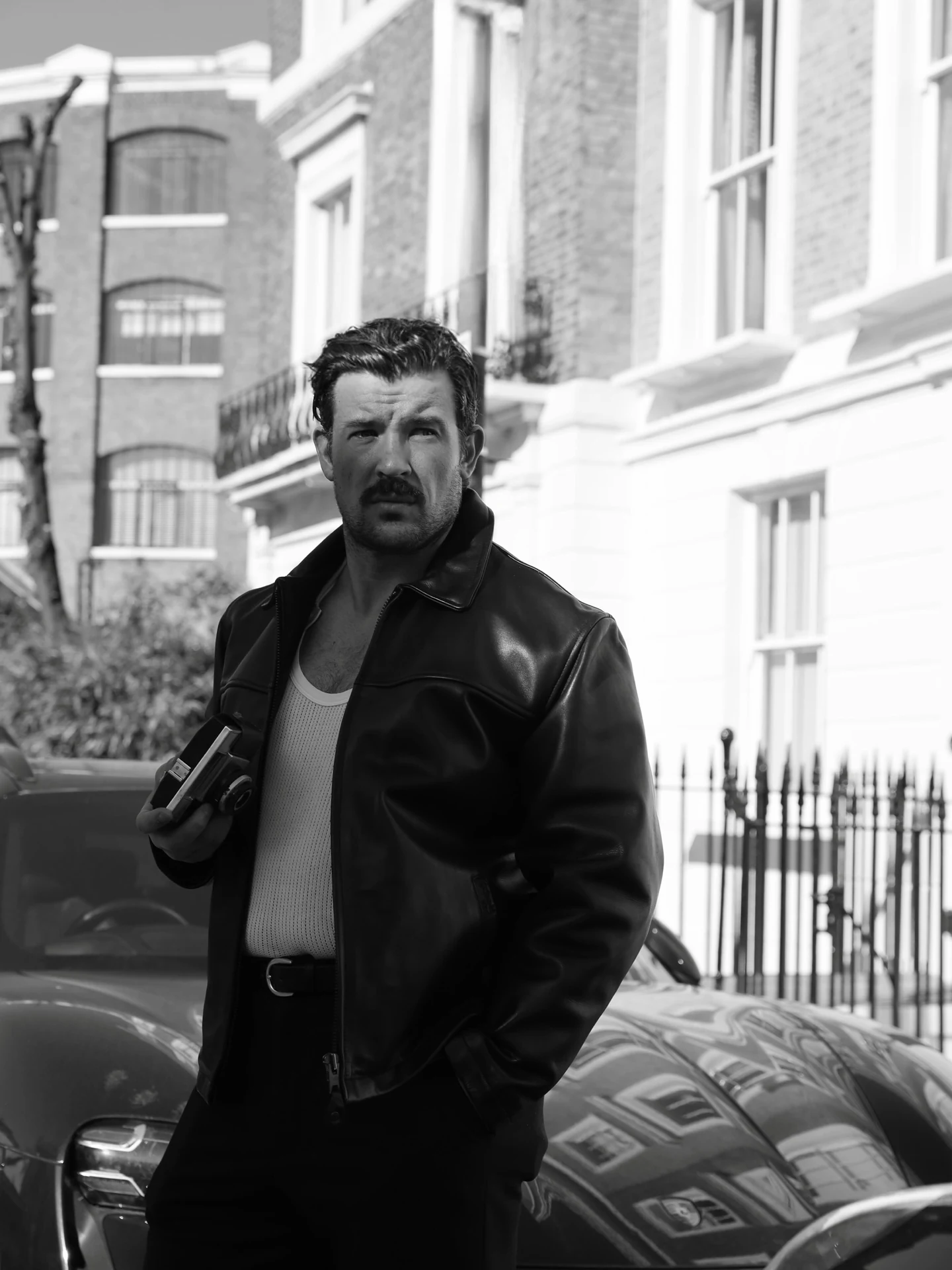 A man in a leather jacket beside London townhouses, photographed in black and white by Paris Golding