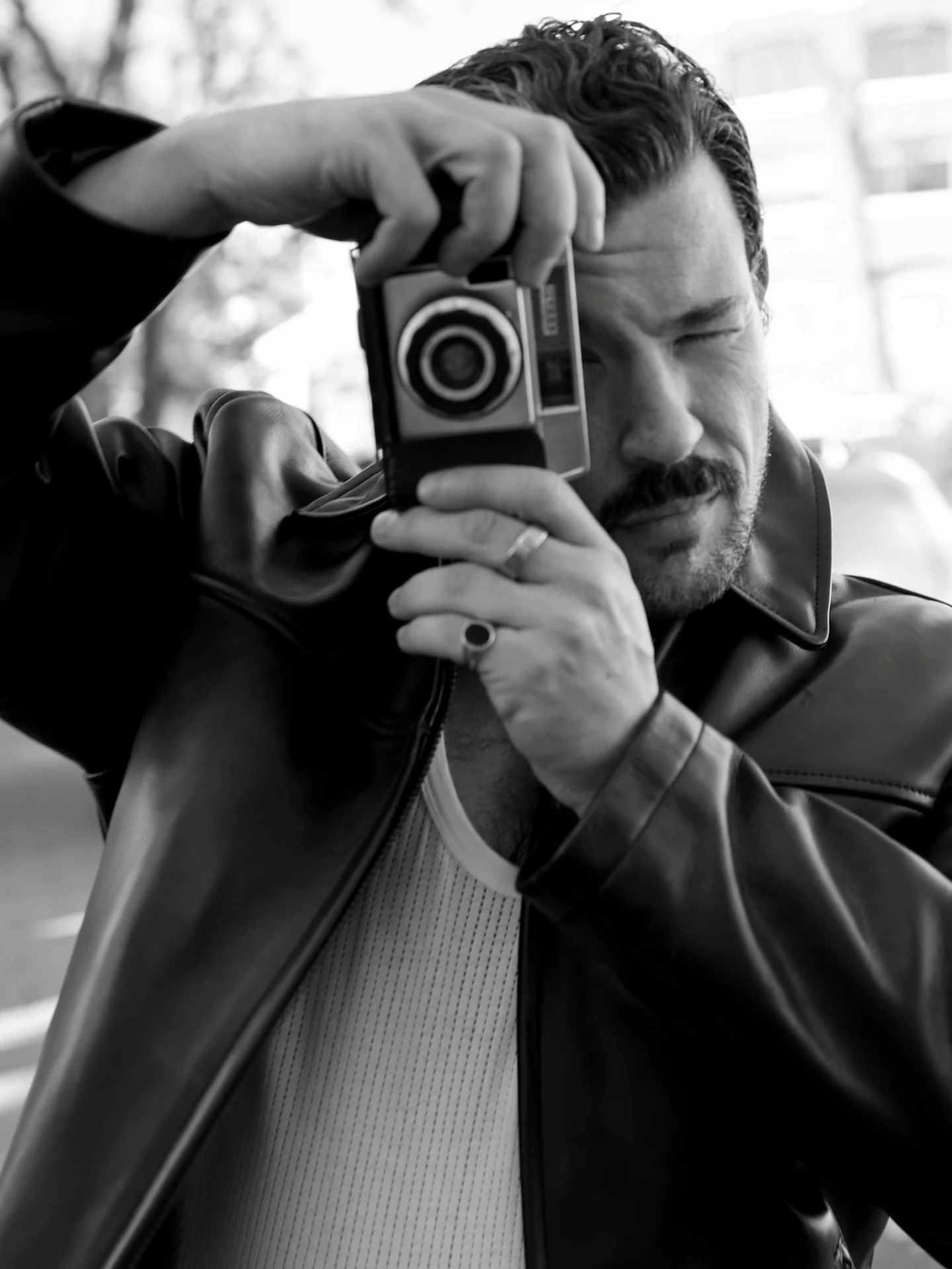 Black-and-white close portrait of a man looking through a camera viewfinder.